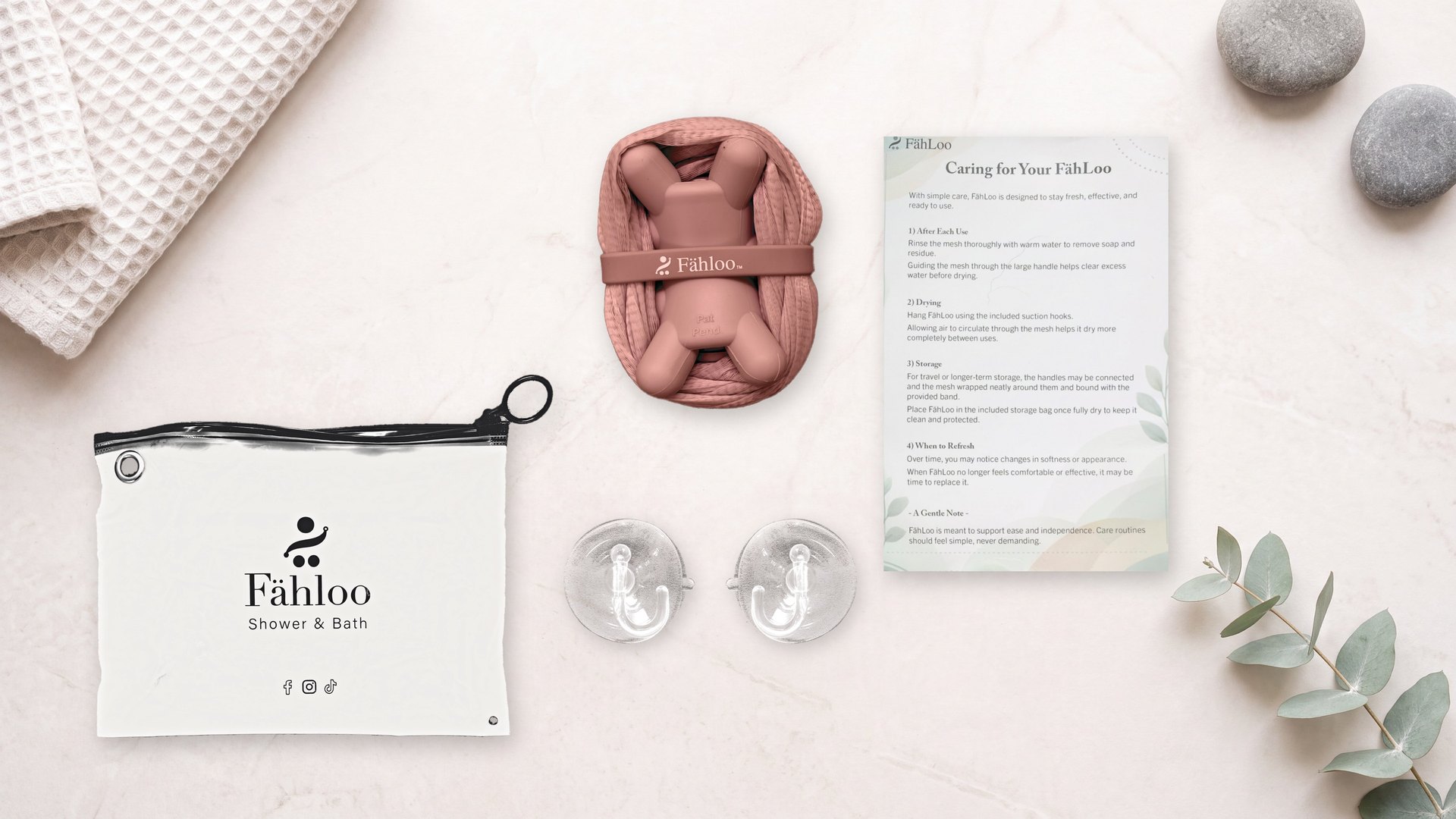 Fähloo package contents on a marble spa surface: the waterproof Fähloo zip bag, a Desert Rose Fähloo bundled as a washcloth, two clear suction hooks, and the Caring for Your Fähloo card